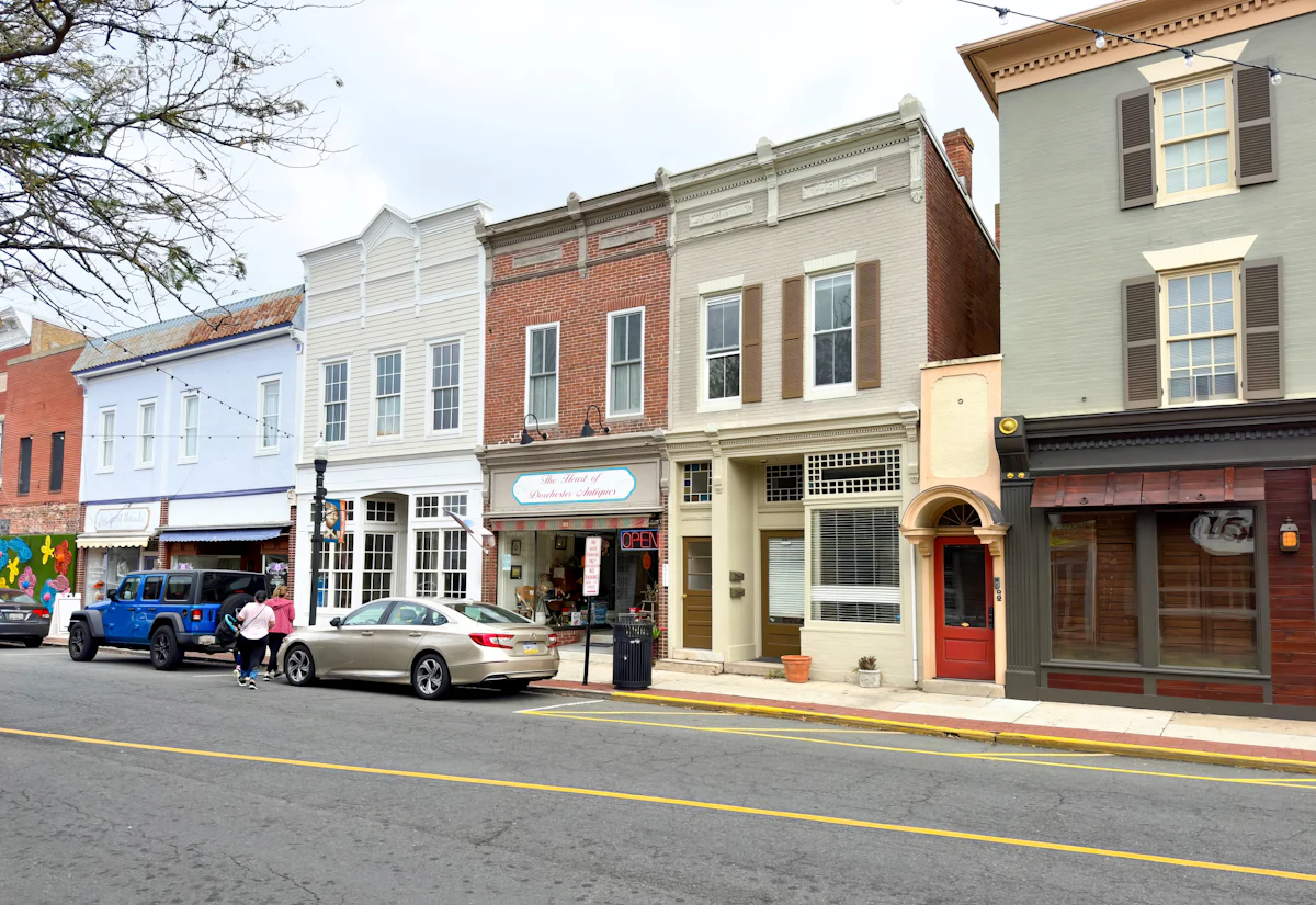 A row of two-storey storefronts on a small-town main street: white clapboard, red brick and grey fronts, an antiques shop with a lit OPEN sign, cars parked at the curb.